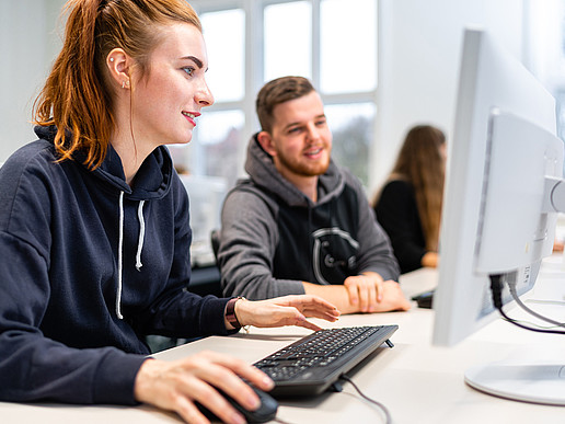 Eine Studentin und ein Student sitzen im Computerkabinett vor einem PC-Bildschirm und wenden diesem ihren Blick zu.