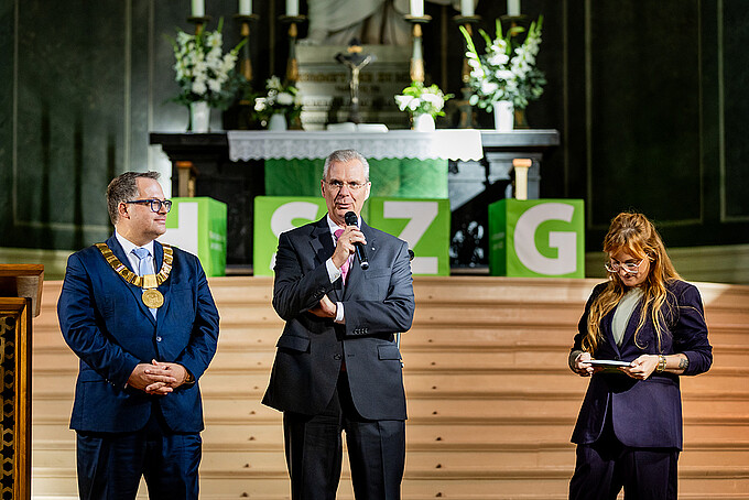 Three people are standing in front of the green HSZG cubes in the chancel during a discussion about the Germany Scholarships.
