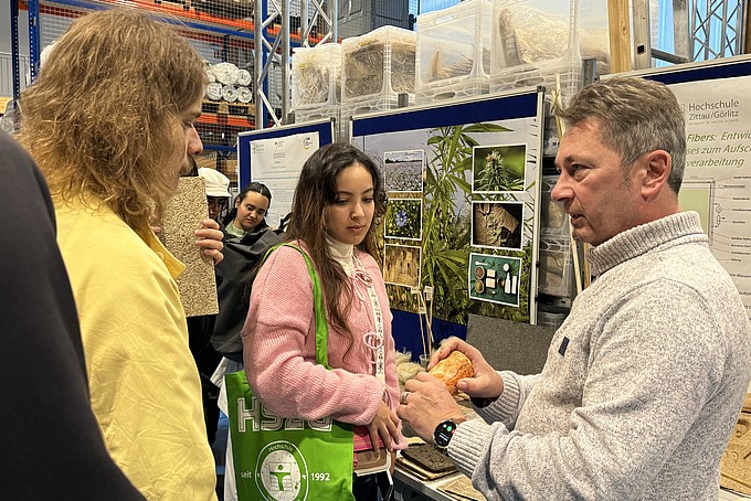 Matthias Tirsch steht in der LANDER³ Halle der HSZG und zeigt einer Gruppe von Besucher verschiedene nachhaltige Materialien. Im Hintergrund sind Informationsposter und gelagerte Materialien zu sehen. Die Teilnehmenden hören aufmerksam zu und betrachten die präsentierten Objekte, während Tirsch die innovativen Anwendungsmöglichkeiten und den Beitrag dieser Forschung zum Umweltschutz erklärt.