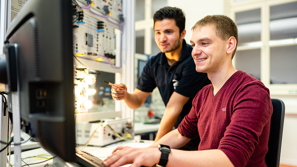 Two students in the electrical engineering lab, one sitting at the computer, the other standing behind it and pointing at the screen.