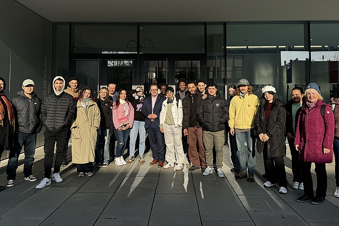 Eine Gruppe von internationalen Studieninteressierten des Rahn Studienkollegs Leipzig posiert gemeinsam mit dem Rektor der HSZG vor dem Eingangsbereich der Hochschule für ein Gruppenfoto. Alle Personen stehen dicht beieinander und lächeln in die Kamera, während die Nachmittagssonne den Eingang des Gebäudes beleuchtet.