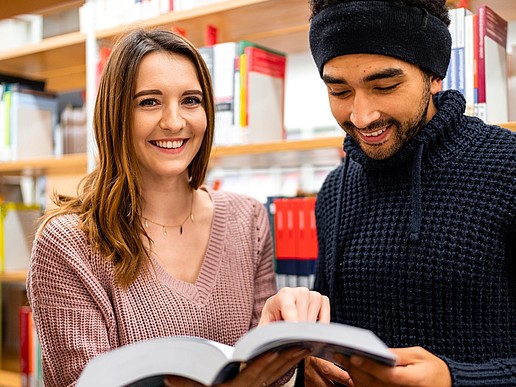 Two students, a young woman and a young man from a migrant background, are standing in the library looking at a book.