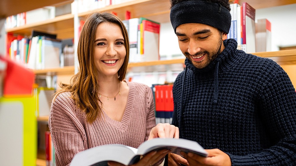 Zwei Studierende, eine junge Frau und ein junger Mann mit Migrationshintergrund stehen in der Bibliothek und schauen sich ein Buch an.