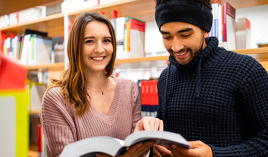 Foto: Tobias Ritz Zwei Studierende, eine junge Frau und ein junger Mann mit Migrationshintergrund stehen in der Bibliothek und schauen sich ein Buch an.