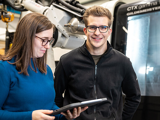 Two students are standing at a machine with a mechanical gripper arm. The student is looking at a tablet.