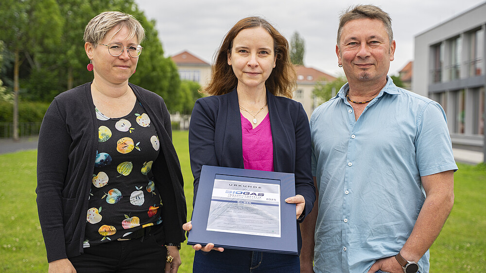 Dr. Marzena Poraj-Kobielska, Dr. Judit Harsányi and Dipl.-Ing. Matthias Tirsch stand on a meadow on the Zittau campus axis. Dr. Harsanyi holds the certificate in her hands.