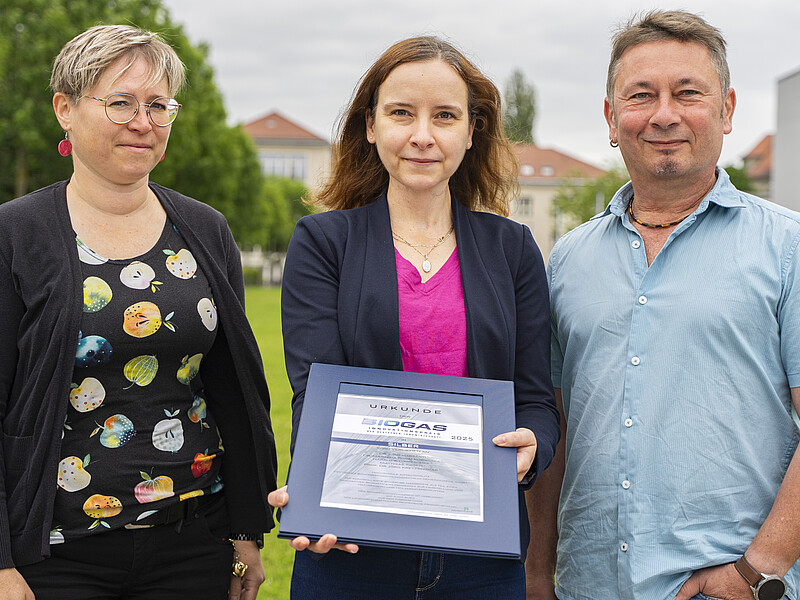 Dr. Marzena Poraj-Kobielska, Dr. Judit Harsányi and Dipl.-Ing. Matthias Tirsch stand on a meadow on the Zittau campus axis. Dr. Harsanyi holds the certificate in her hands.