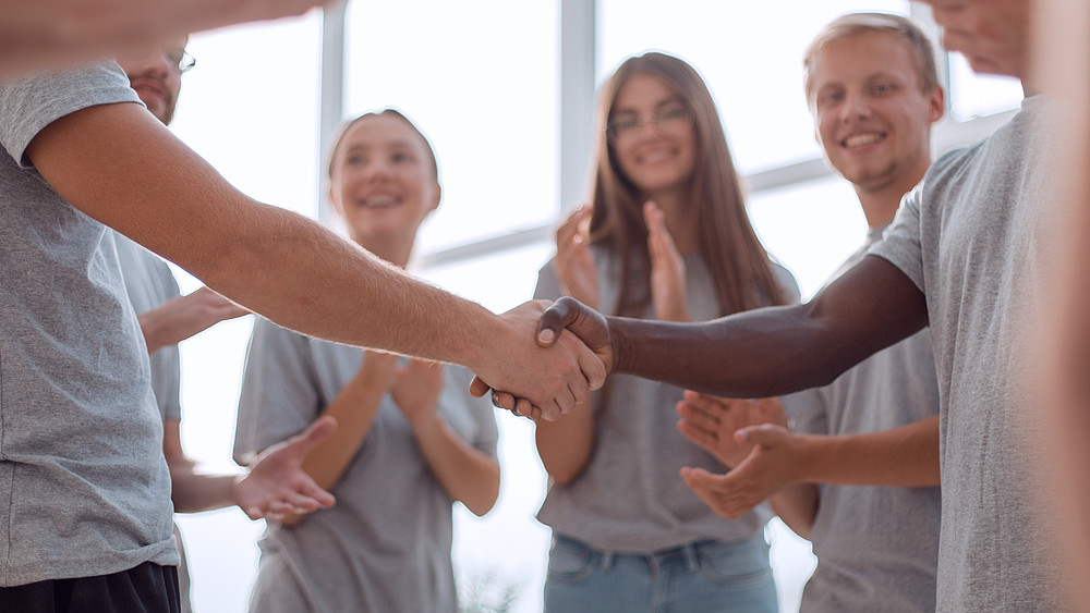 Two group members shake hands, young students stand by and applaud.