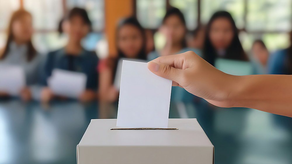 Young people are sitting in the lecture hall in the background, in front of them you can see a hand inserting a white piece of paper into a ballot box.