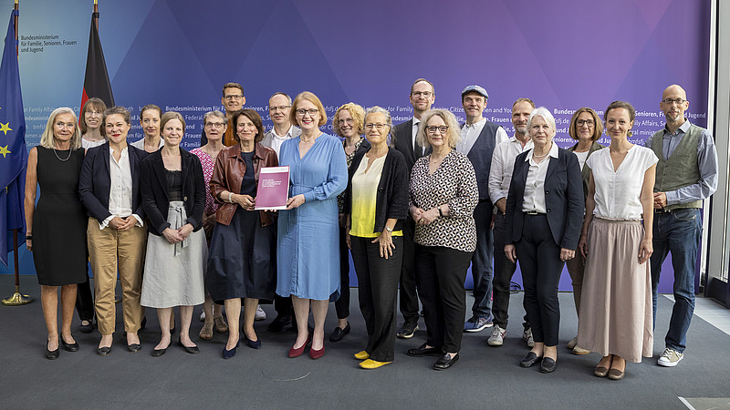 Beiratsmitglieder und die Familienministerin vor einer Pressewand im Bundestag stehen für ein Gruppenbild nebeneinander. Die Ministerin hält den Bericht in der Hand.