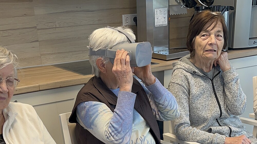 An old lady is wearing VR glasses, two old ladies are sitting to her left and right.