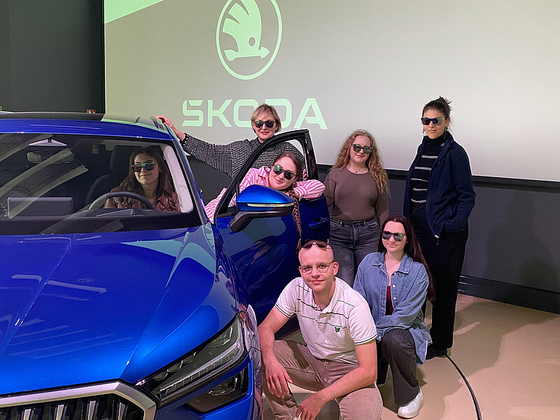 Several students stand and squat next to a blue Skoda.