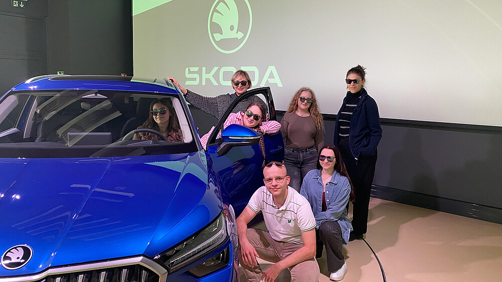 Several students stand and squat next to a blue Skoda.