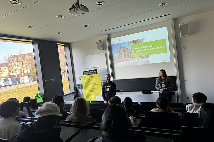 Robert Viertel und Inna Hauf stehen vorne in einem Hörsaal und sprechen zu den internationalen Studieninteressierten des Rahn Studienkollegs Leipzig. Im Hintergrund ist eine Präsentation mit dem Titel „Herzlich Willkommen an der Hochschule Zittau/Görlitz“ zu sehen. Die Zuhörer sitzen in Reihen und folgen aufmerksam der Begrüßung.