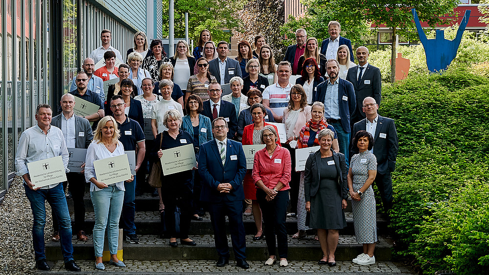 A group of university members and cooperation partners from the care sector stand next to the Blue Box on the Görlitz campus.