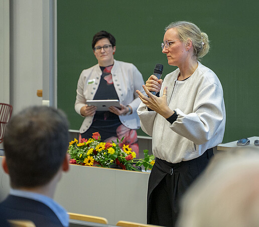 Two women stand in front of the audience, one speaks into a hand-held microphone.