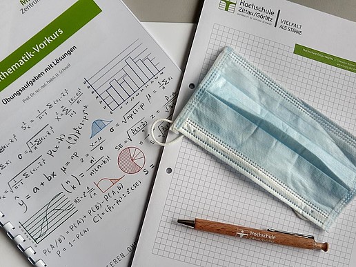 On the left side you can see the title page of the exercises for the math pre-course, on the right side a checkered writing pad with the university logo. On the pad is a typical medical disposable mouth and nose protector and a wooden ballpoint pen with the university logo.
