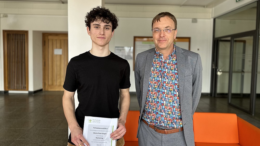 Tom Saugel with certificate in his hands and Prof. Kroschel standing in the hallway of a university building.