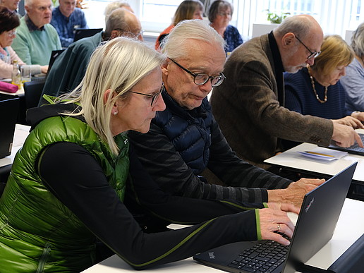 A senior citizen sits in the seminar room surrounded by other senior citizens and looks at a laptop.