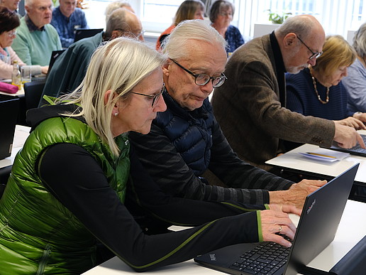 Eine Seniorin und ein Senior sitzen im Seminarraum umgeben von anderen Seniorinnen und Senioren und schauen auf einen Laptop.