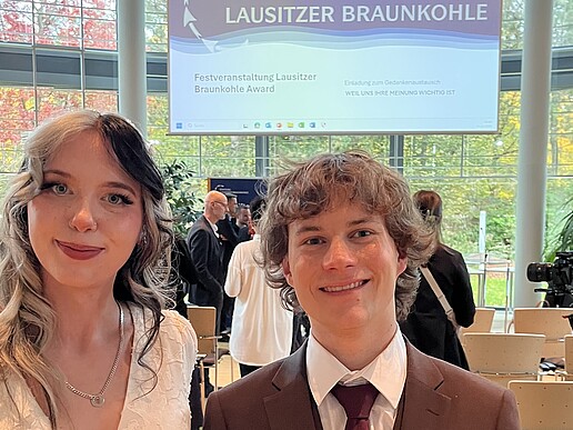 Zwei Studierende stehen in einem Veranstaltungssaal und lächeln in die Kamera.