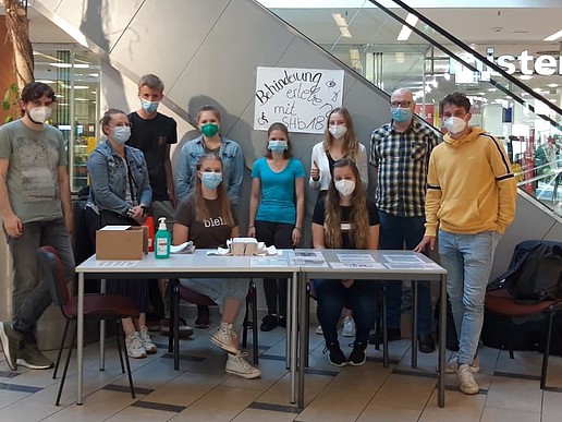 A group of students and a lecturer stand next to an escalator in a shopping center around a table on which various materials for making disability tangible are laid out.