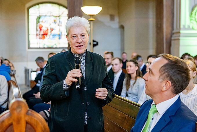 Ellen Leuschner stands between the pews with a microphone and speaks to the audience.