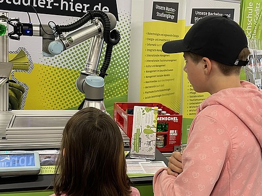 A robotic arm uncorks a bottle of lemonade. Two children watch.