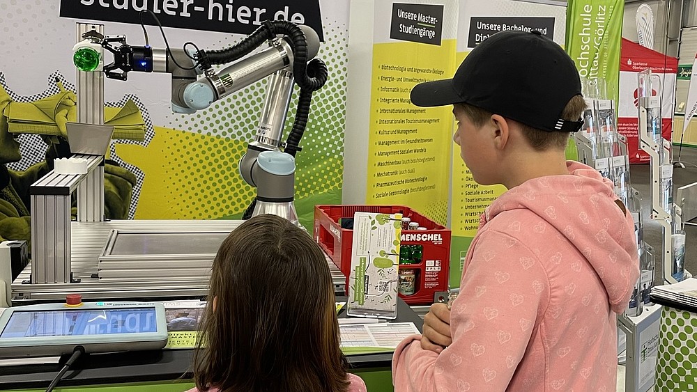 Ein Roboterarm entkorkt eine Flasche Limonade. Zwei Kinder schauen zu.