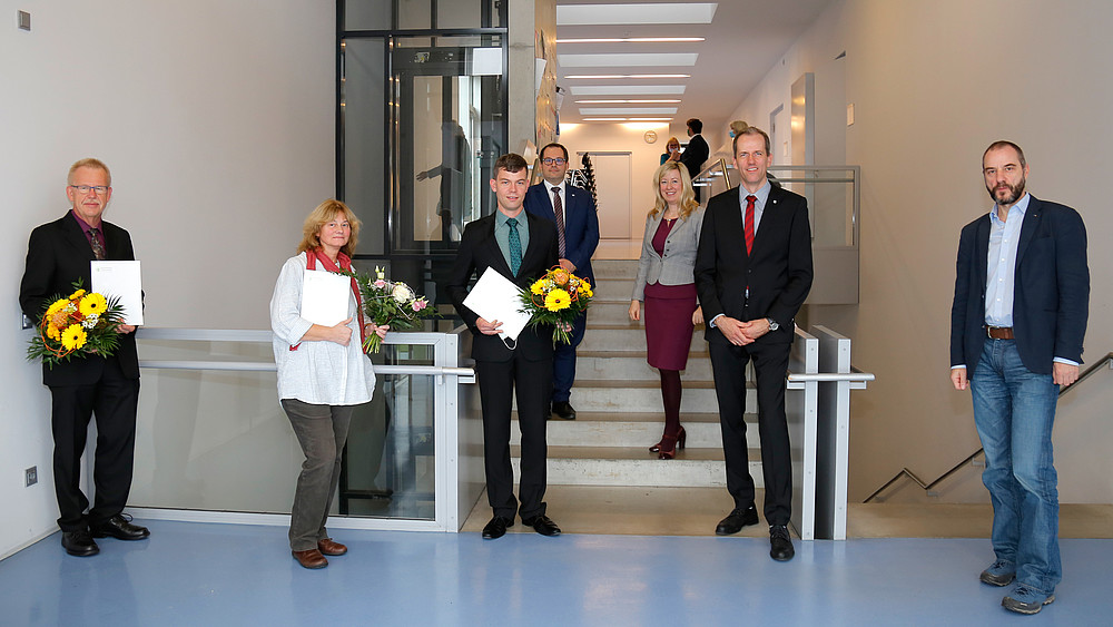 Eine Gruppe von Personen stehen verteilt im Treppenhaus. Drei halten Blumensträuße und Urkunden in der Hand. Alle lächeln in die Kamera.