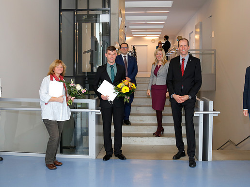 A group of people are standing in the stairwell. Three are holding bouquets of flowers and certificates. They are all smiling at the camera.