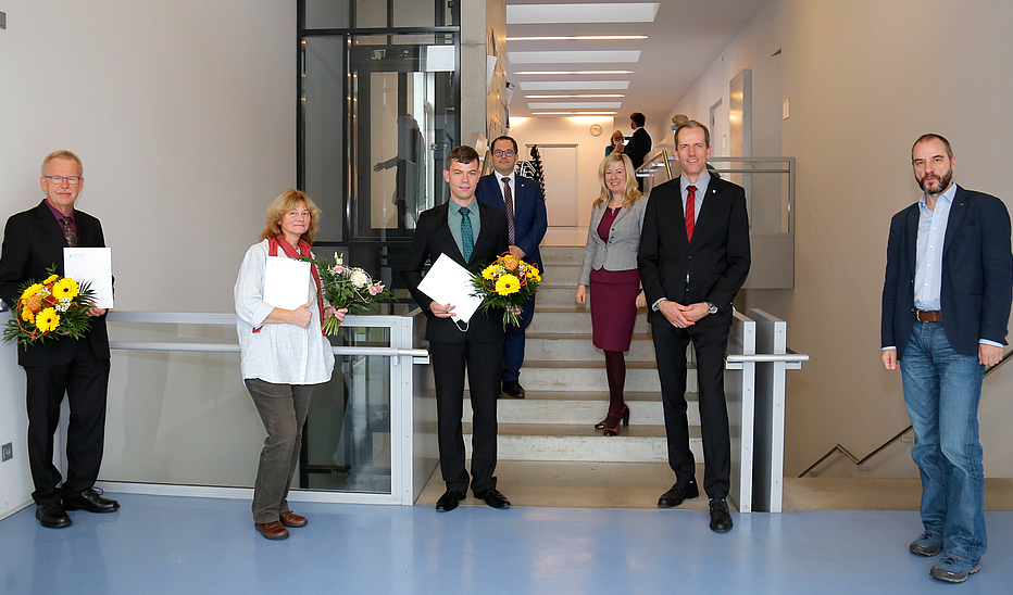 Foto: Jens Freudenberg Eine Gruppe von Personen stehen verteilt im Treppenhaus. Drei halten Blumensträuße und Urkunden in der Hand. Alle lächeln in die Kamera.
