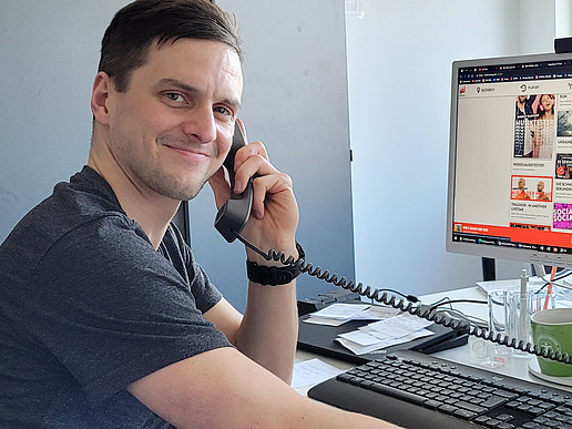 Rico Winkler smiles into the camera, sitting at his desk with a telephone receiver in his hand and the Radio Energy Sachsen website displayed on his PC screen in the background.