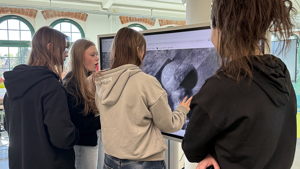 Schülerinnen stehen vor einem Bildschirm, auf dem ein Mondkrater zu sehen ist.