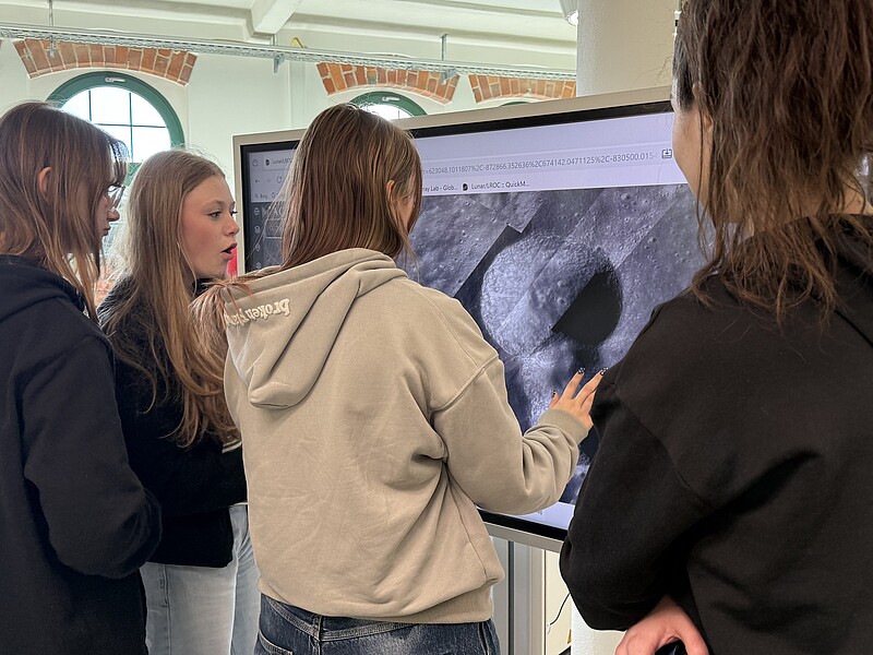 Schülerinnen stehen vor einem Bildschirm, auf dem ein Mondkrater zu sehen ist.