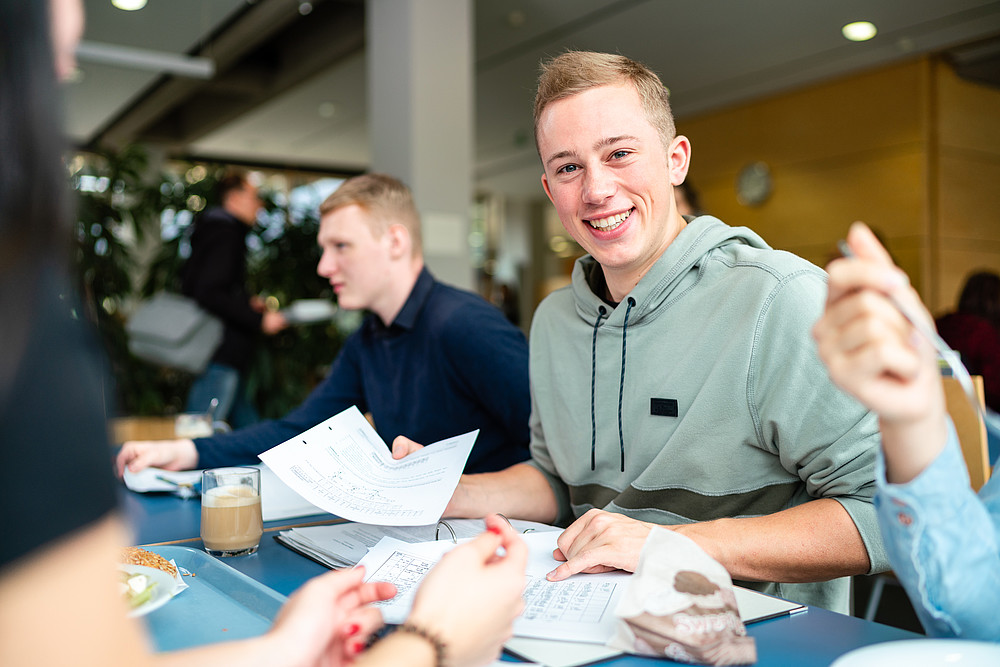 Students at a table in the canteen. Visible are 2 young men with study documents, one of them smiling and looking into the camera.