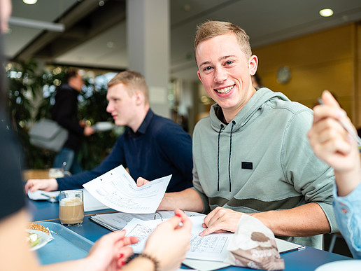 Students at a table in the canteen. Visible are 2 young men with study documents, one of them smiling and looking into the camera.