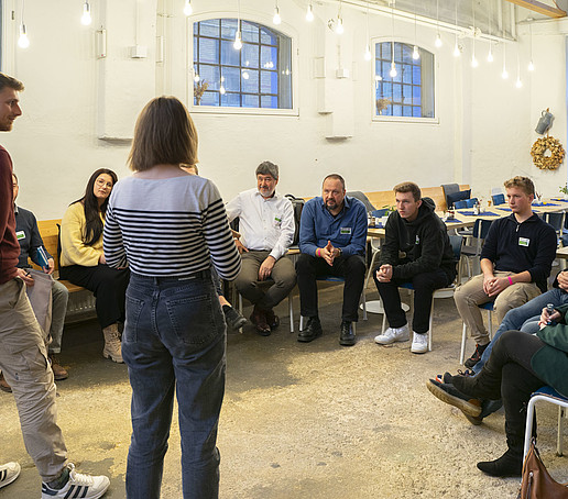 In the room, participants sit on rows of chairs and listen to two standing participants.