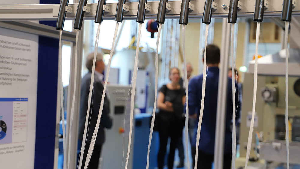 A machine processing natural fibers is in the foreground. People are talking in the background.