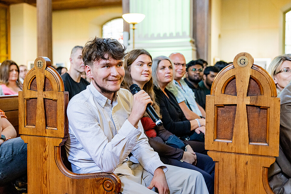 A freshman is sitting in a pew at St. John's Church and speaking into a handheld microphone.