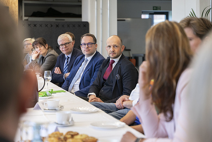 Geschäftsleute sitzen an einem langen Tisch bei einer Besprechung im Vordergrund ist der Hinterkopf einer Person zu sehen, die auf die sprechenden Teilnehmenden blickt.