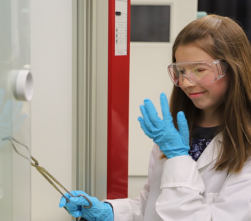 A girl in the laboratory looks at her finger through her safety goggles, fascinated. She is holding a pair of pliers in her other hand.