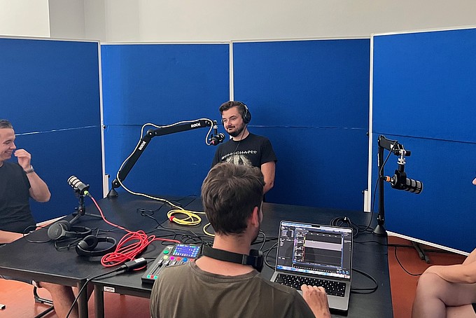 Podcastteilnehmende sitzen am Tisch umgeben von Schallwänden. Das Aufnahmeequipment steht auf dem Tisch.