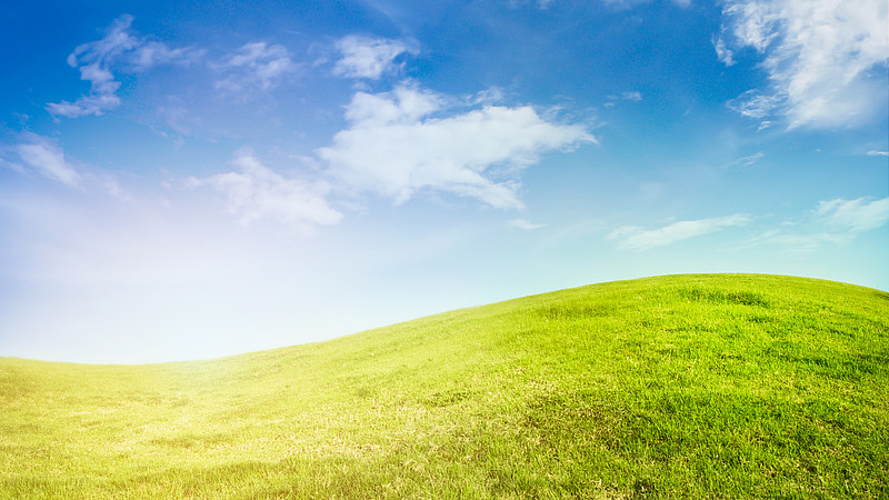 eine grüne Wiese geht in einen begünten Hügel über, darüber ein blauer Himmel mit vereinzelten Wolken