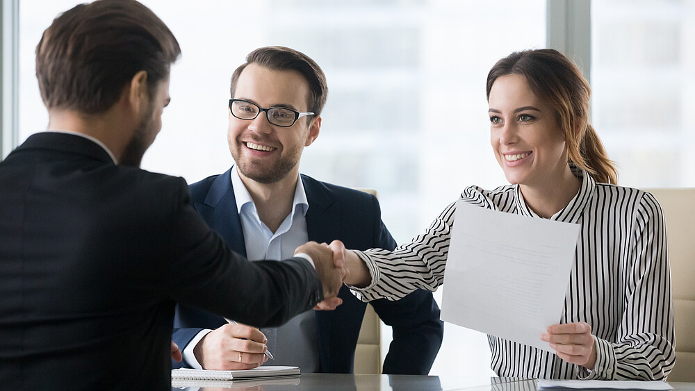 At a table, a man in a suit shakes hands with a woman sitting opposite him, who is holding a piece of paper in her other hand.