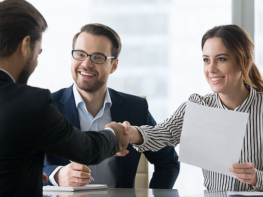 At a table, a man in a suit shakes hands with a woman sitting opposite him, who is holding a piece of paper in her other hand.