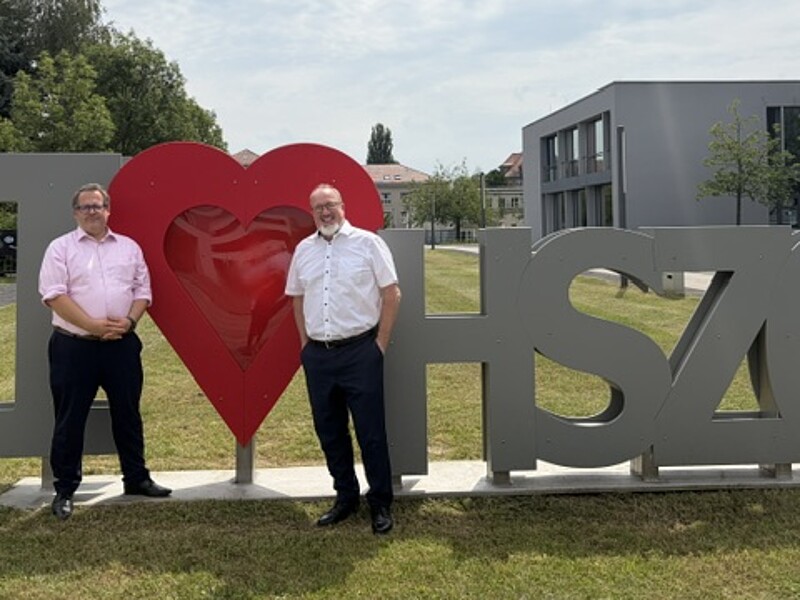 : Prof. Dr.-Ing. Alexander Kratzsch und Prof. Dr.-Ing. Sascha Müller-Feuerstein vor dem I Love HSZG Logo auf dem Campus
