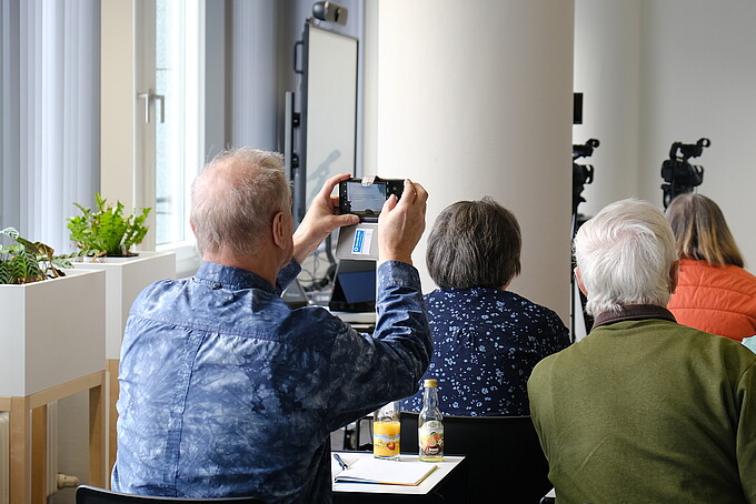 In the seminar room, a senior citizen is filming a lecture with his smartphone.