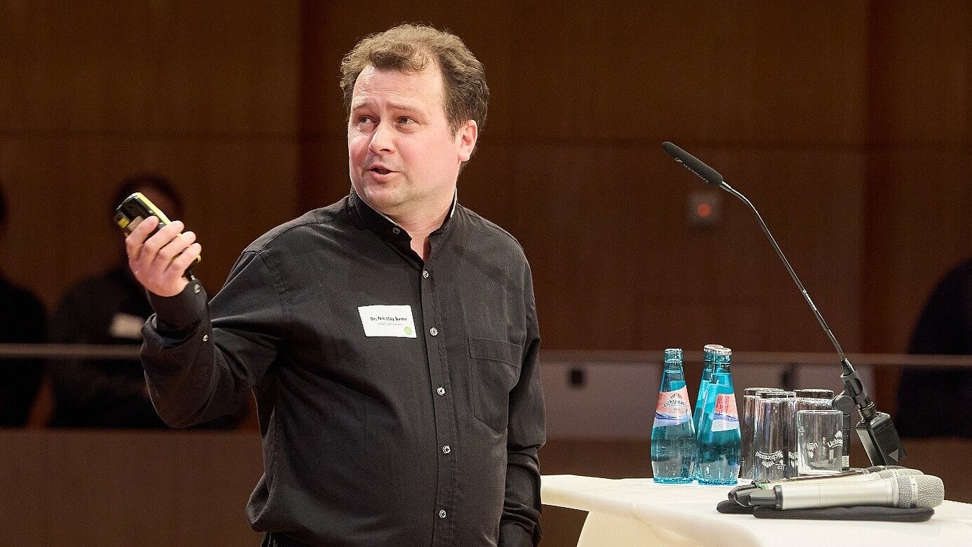 Mr. Belov holds a remote control in his hand and points to the screen behind him.