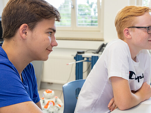 Zwei Schüler sitzen am Tisch in einem Labor und blicken in Richtung Rednerpult.